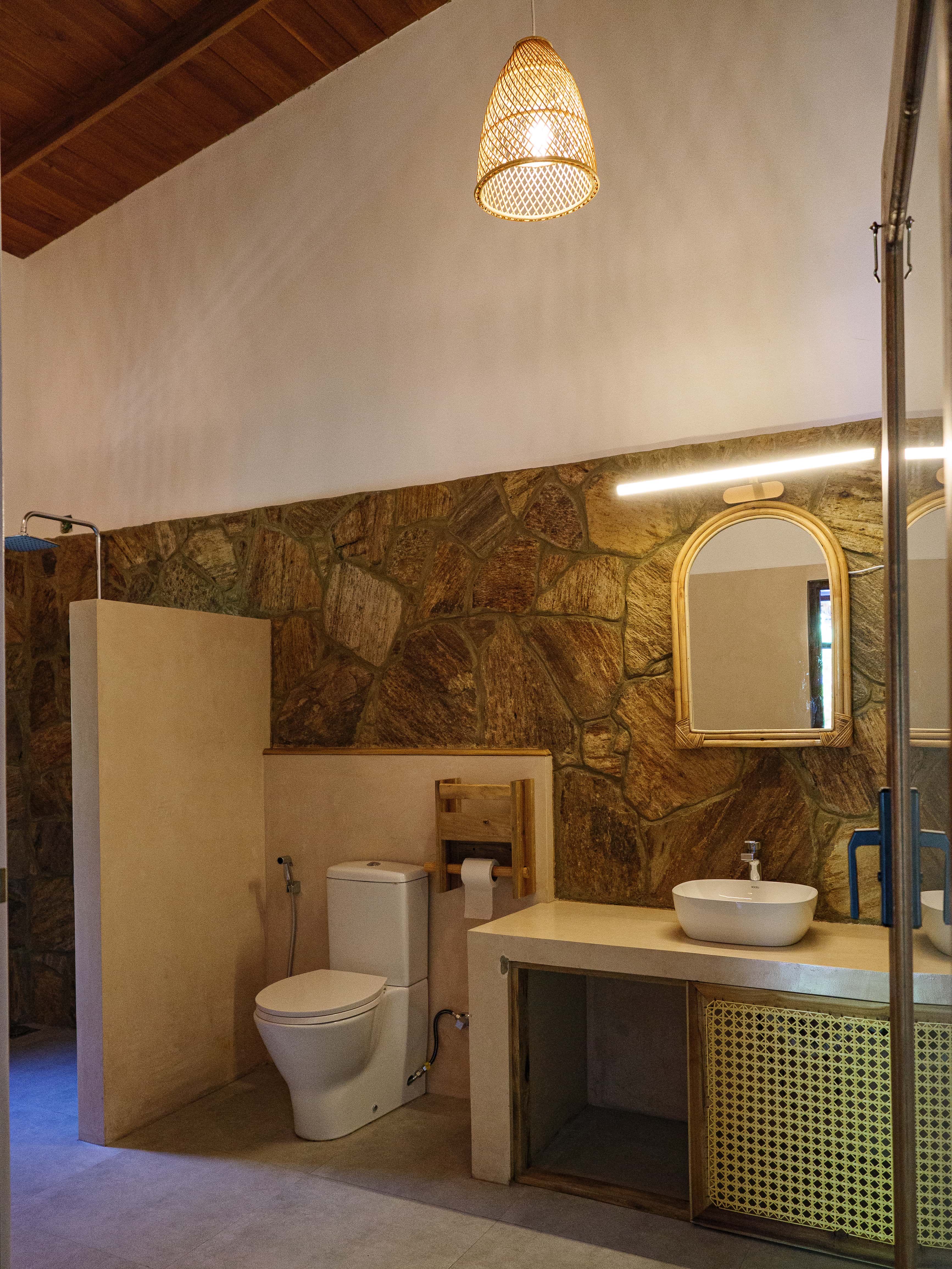 Spacious shared bathroom with natural stone walls, wicker pendant light, and woven rattan vanity at Pine and Palm dorm in Arugam Bay`