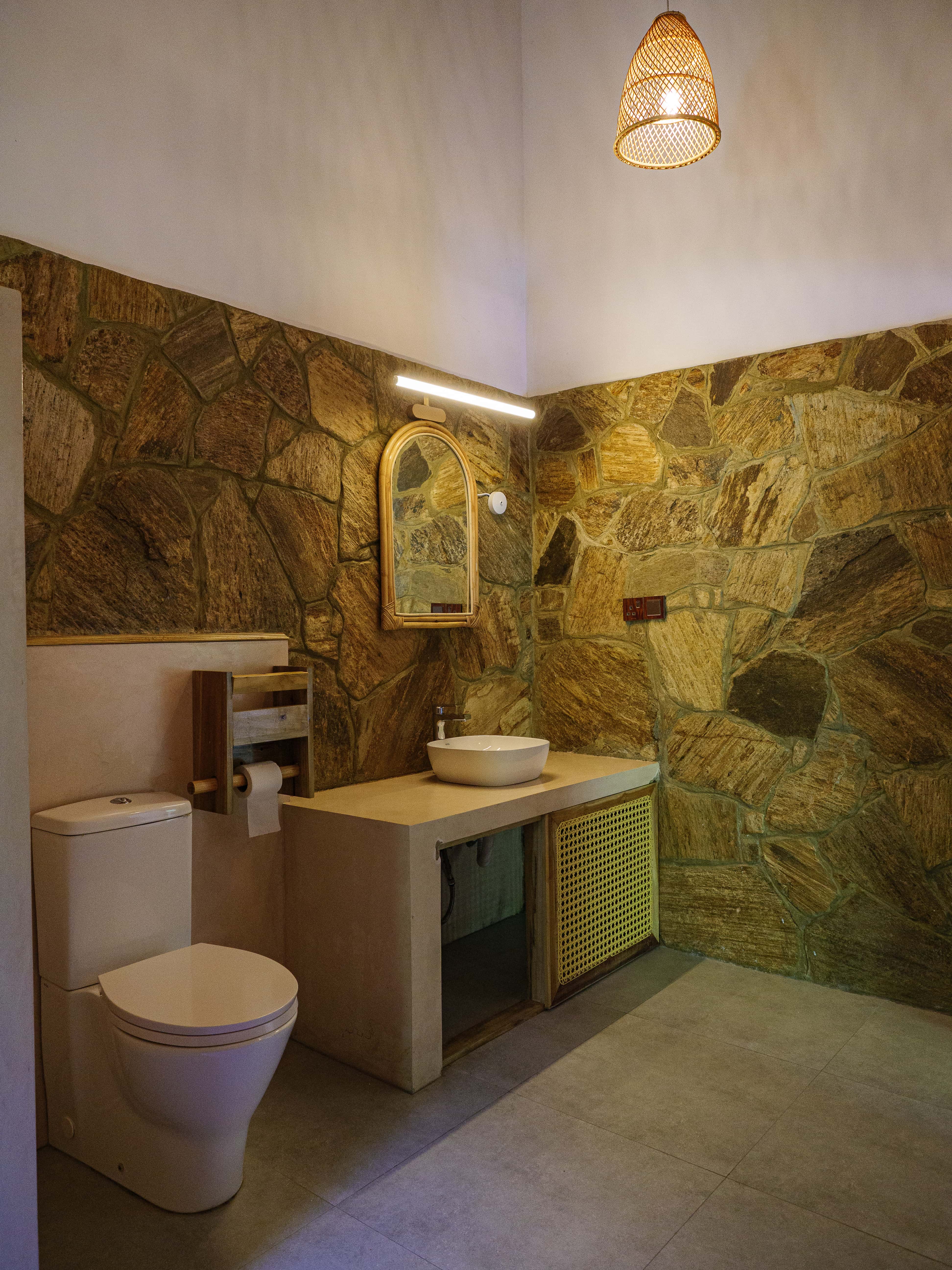 Laid-back boutique bathroom interior with natural stone walls, a woven vanity, and hanging rattan light fixture at Pine and Palm dorm in Arugam Bay