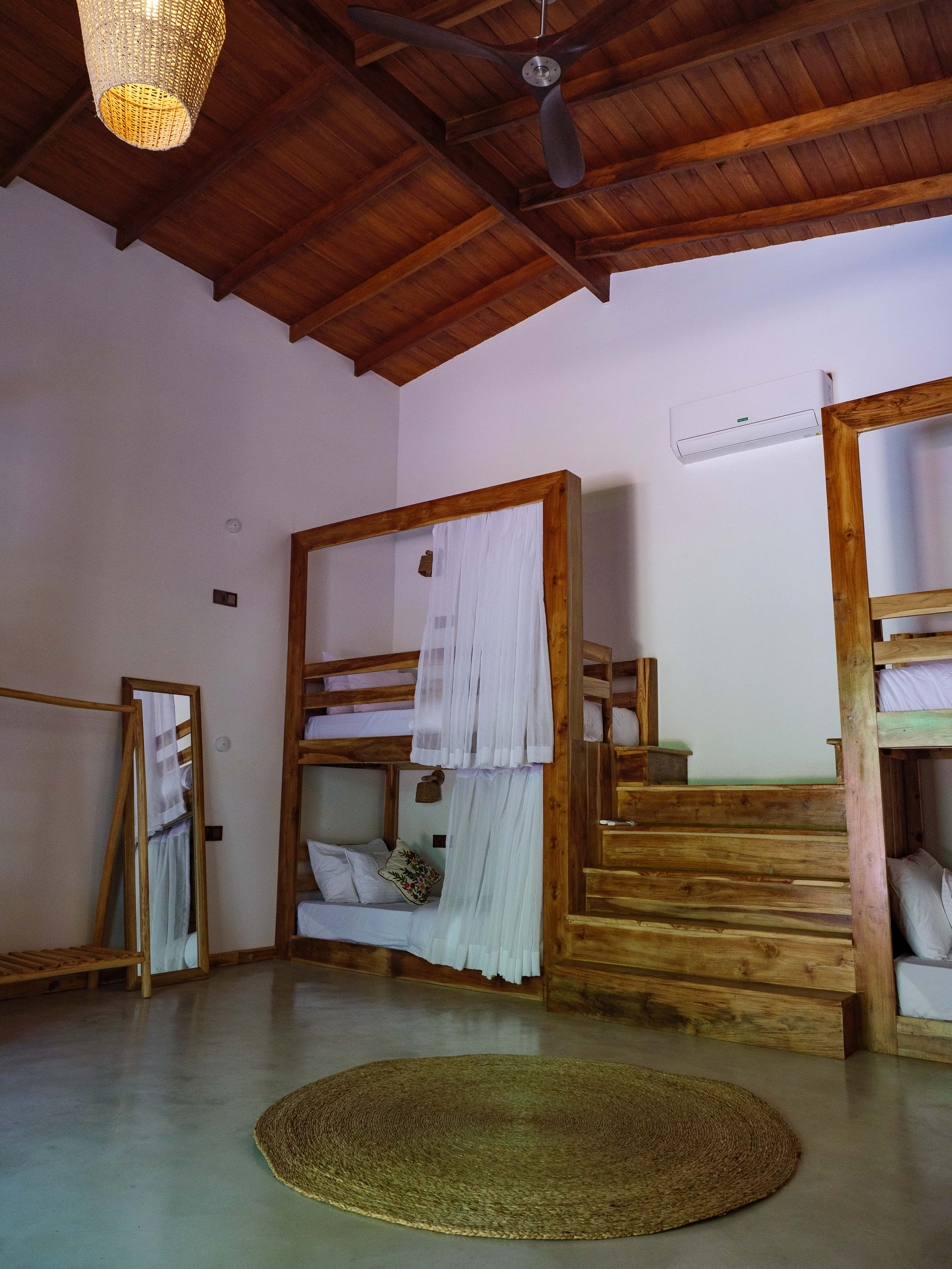 Spacious six-bed dorm room with wooden bunk beds, privacy curtains, high ceilings, and a woven jute rug at Pine and Palm in Arugam Bay