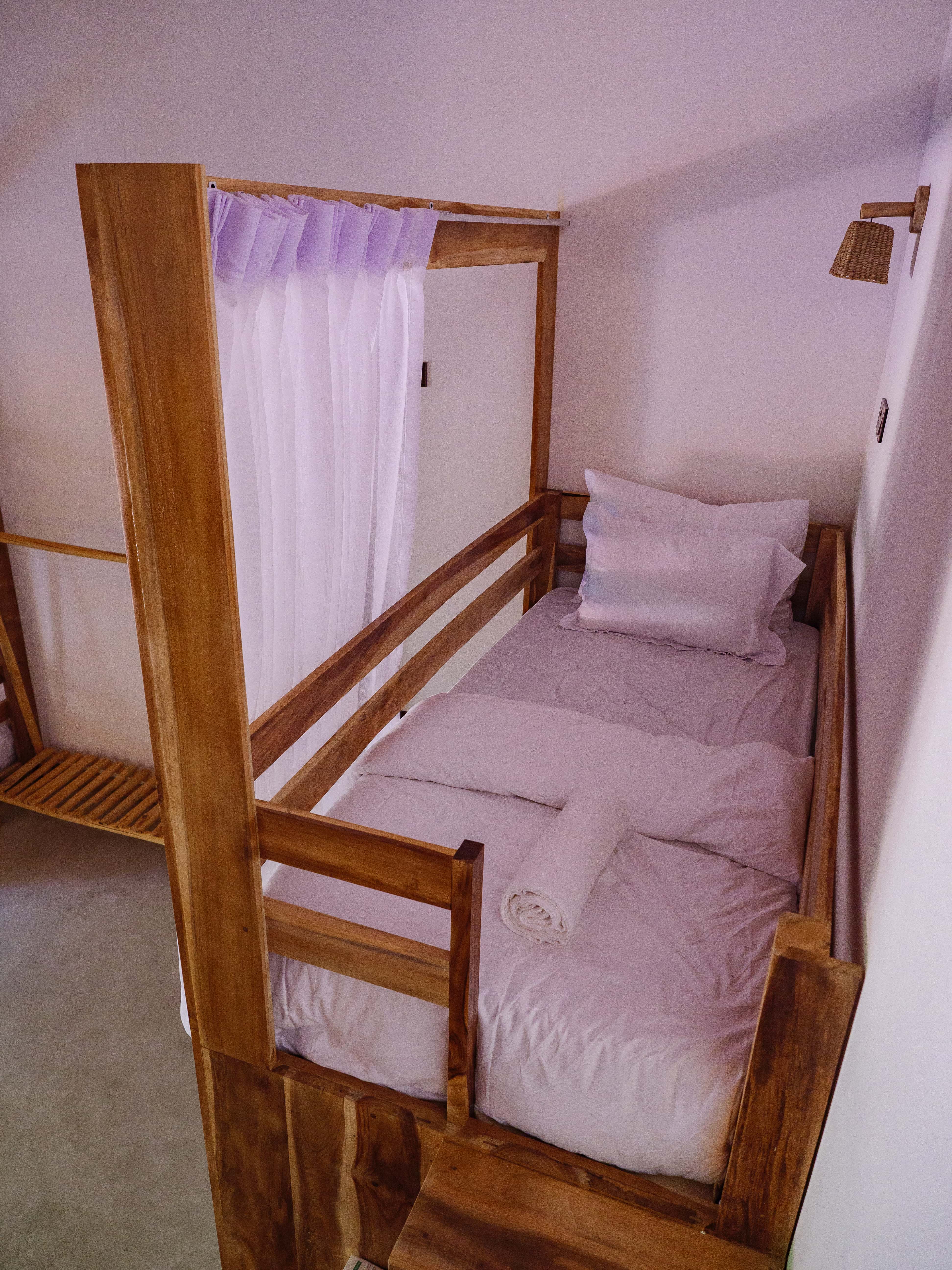 Top bunk bed with fresh white linens, a rolled towel, and privacy curtains in the shared dorm room at Pine and Palm in Arugam Bay