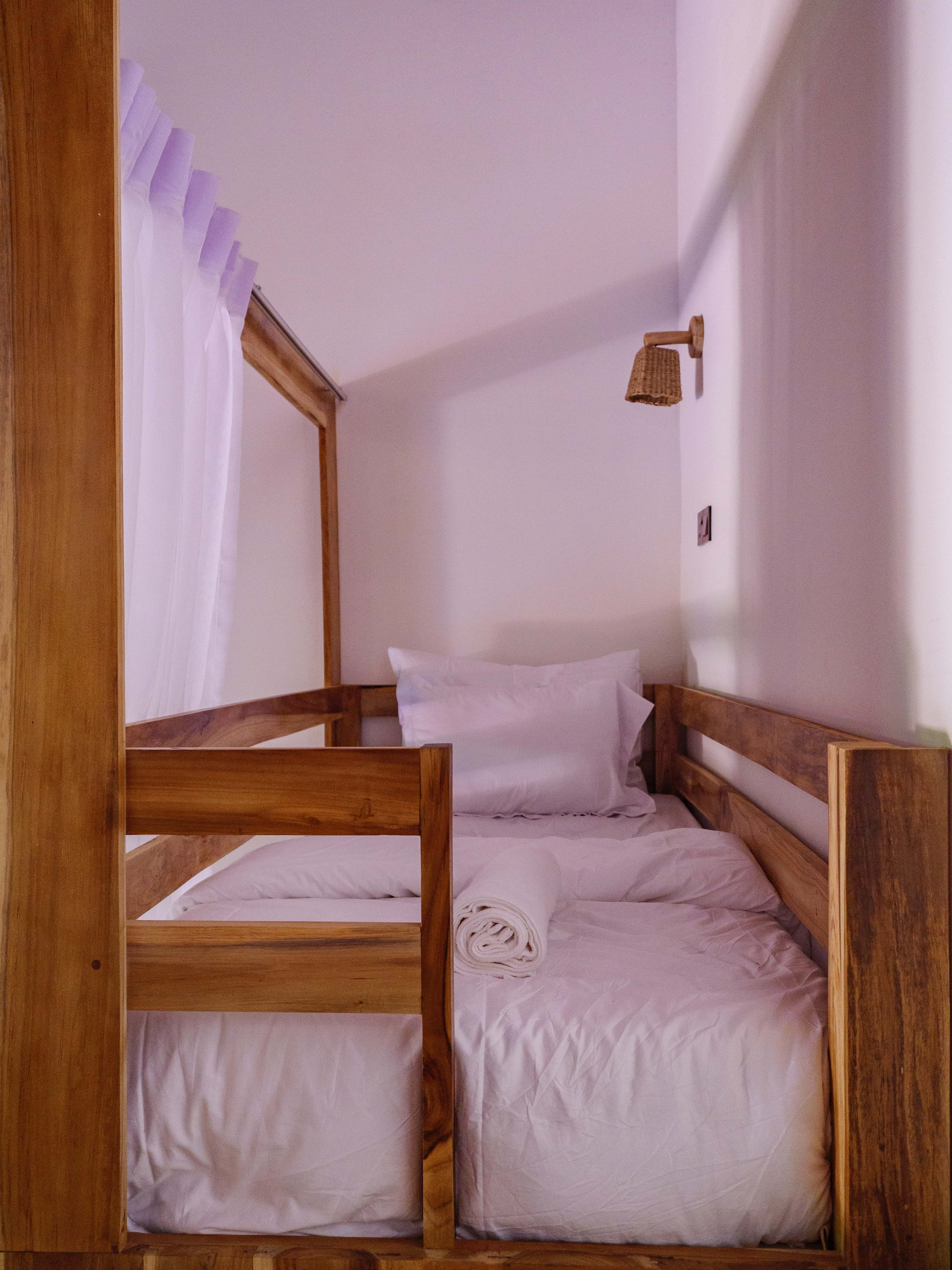 Cozy top bunk bed with white bedding, a rolled towel, privacy curtains, and a woven wall reading lamp in the shared dorm at Pine and Palm in Arugam Bay