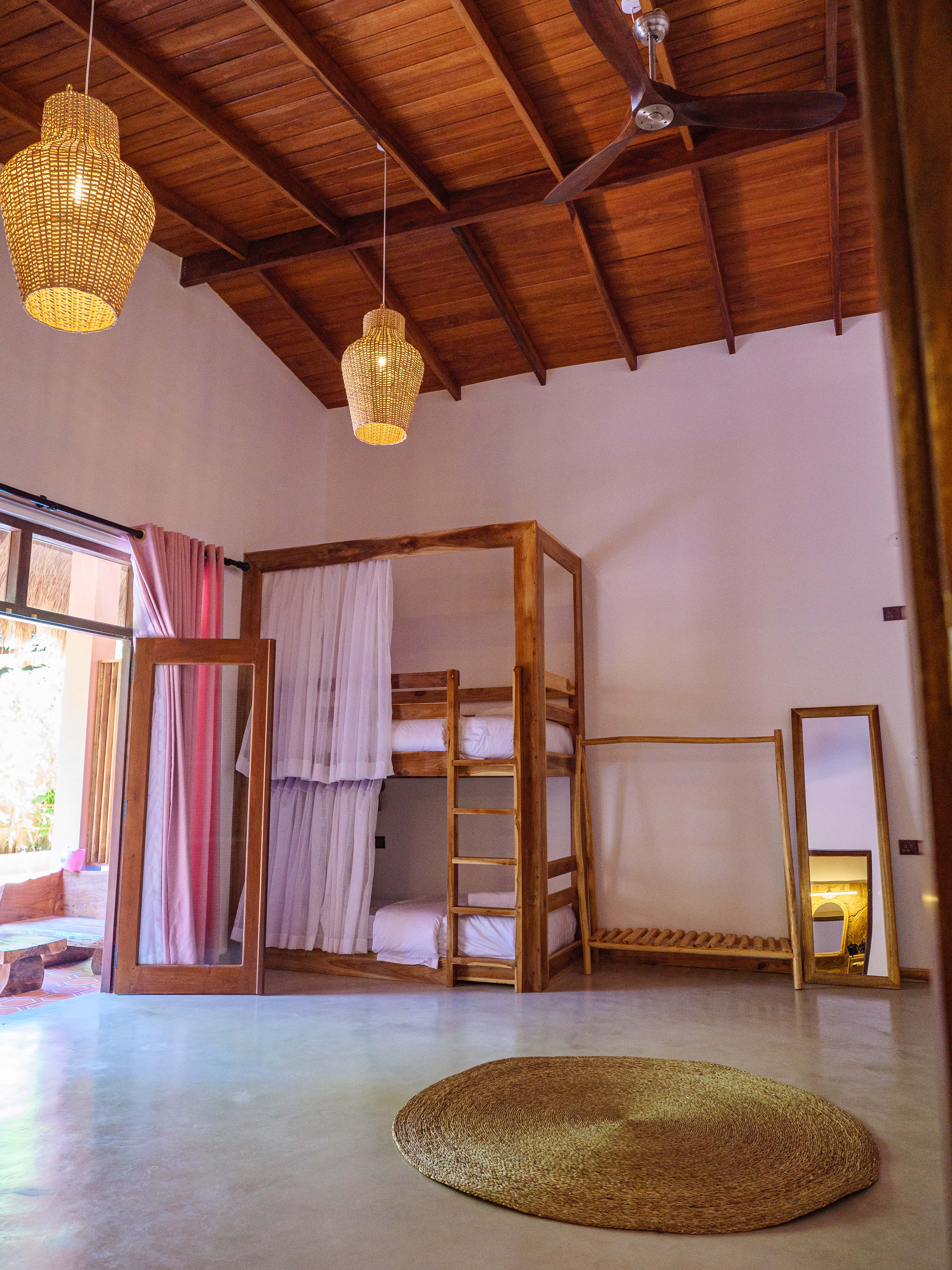 Bright shared dorm room with wooden bunk beds, privacy curtains, high ceilings with wicker pendant lights, and a round jute rug at Pine and Palm in Arugam Bay
