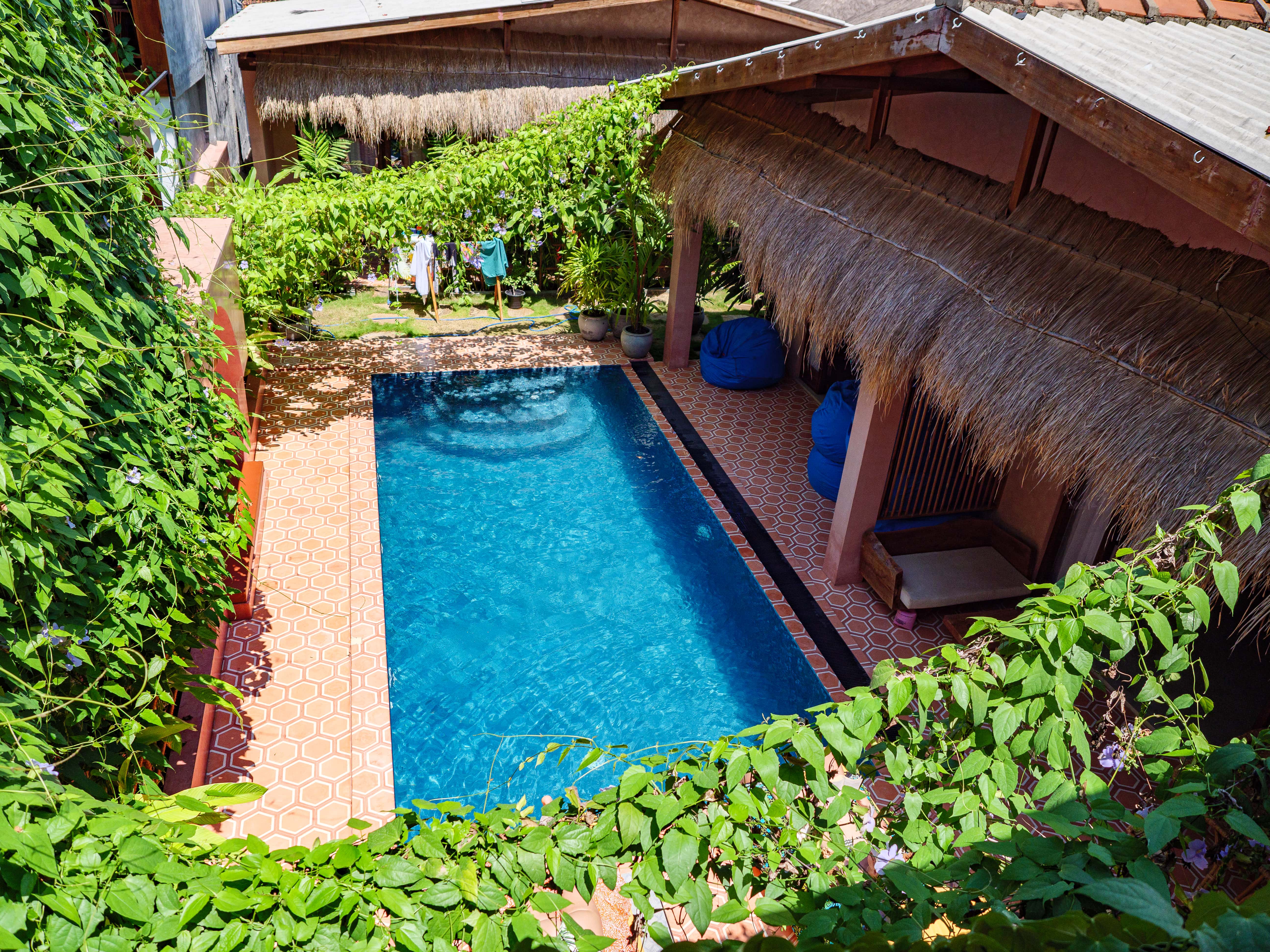 High-angle view of the outdoor swimming pool surrounded by tiled courtyard, blue bean bags, and thatched roof bungalows at Pine and Palm in Arugam Bay
