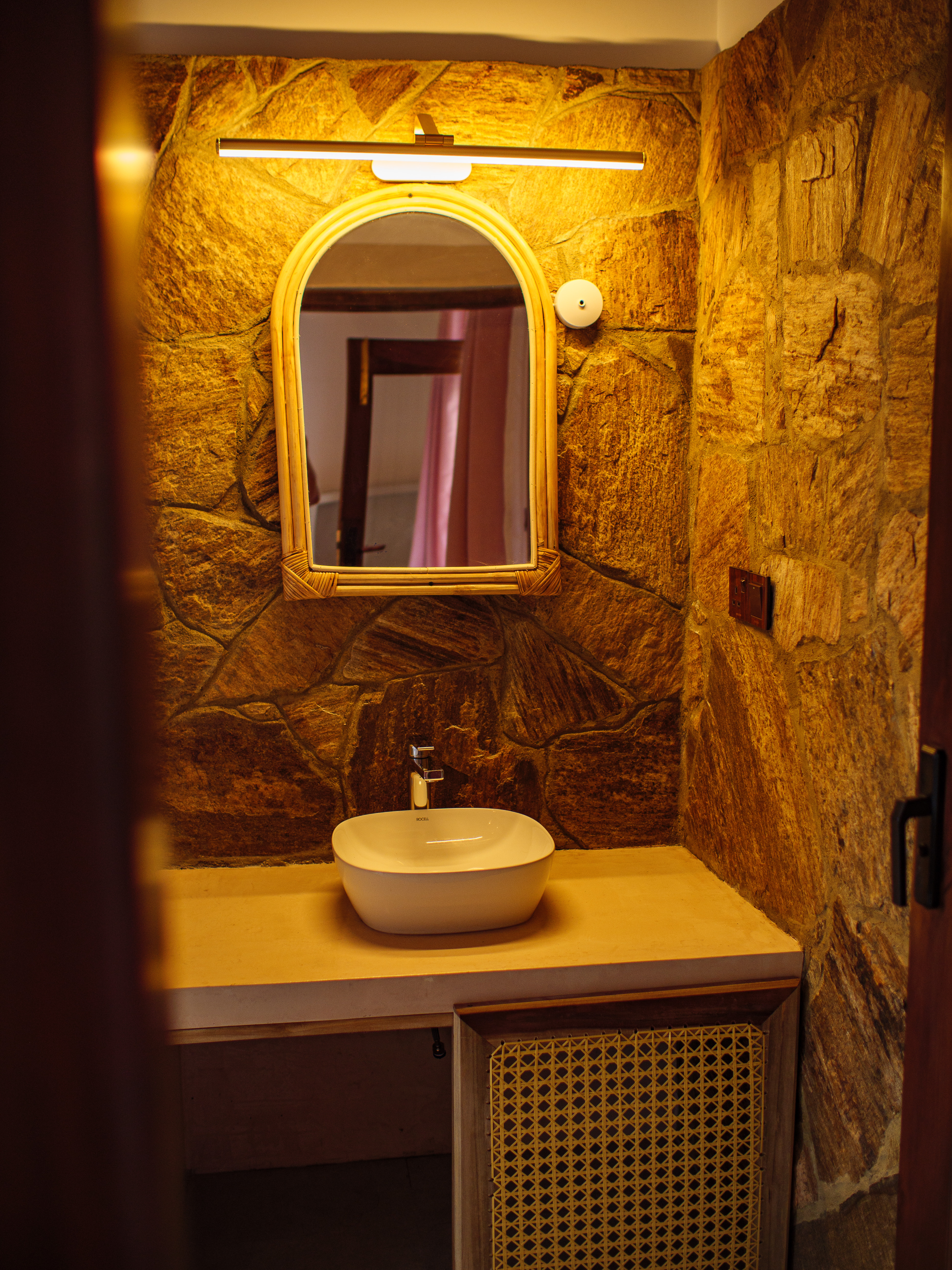 Rustic stone wall bathroom with vessel sink, arched rattan mirror, and warm lighting at Pine and Palm boutique stay in Arugam Bay